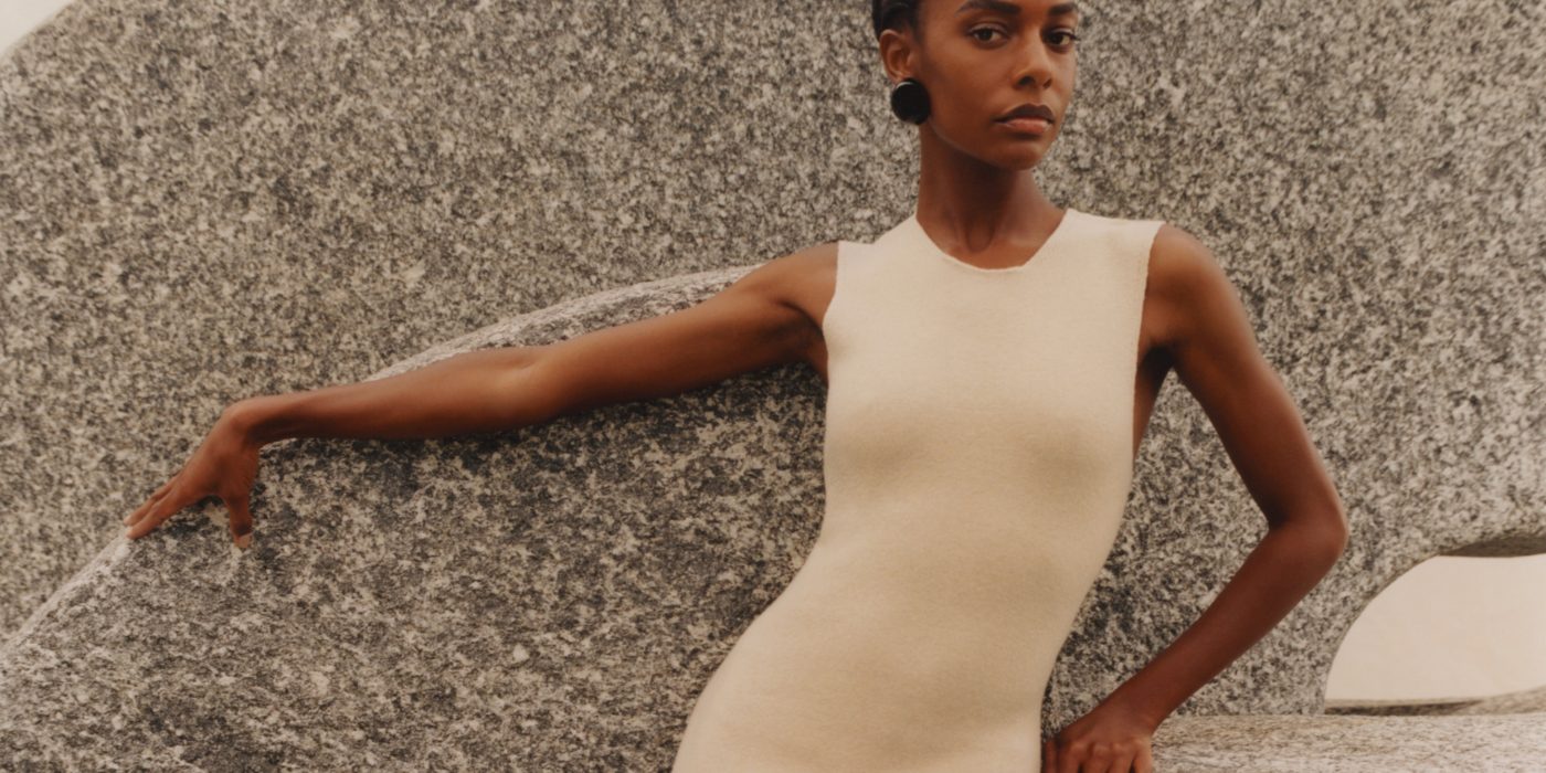 A woman wearing a sleeveless beige dress from Giorgio Armani's SS22 collection poses against a rock wall.