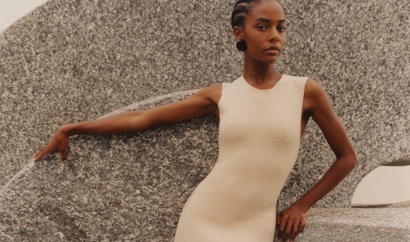 A woman wearing a sleeveless beige dress from Giorgio Armani's SS22 collection poses against a rock wall.
