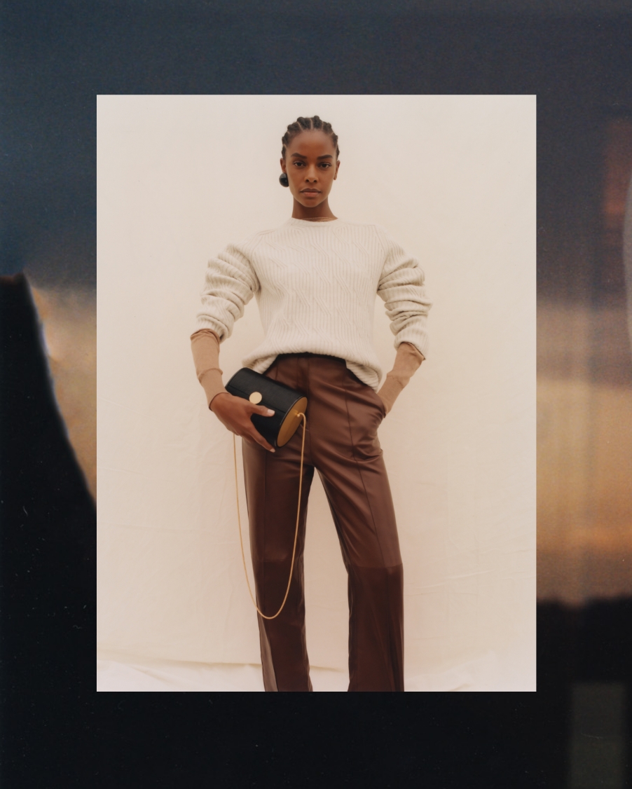 A woman wearing a cream sweater and brown pants holds a black purse from Giorgio Armani's SS22 Collection