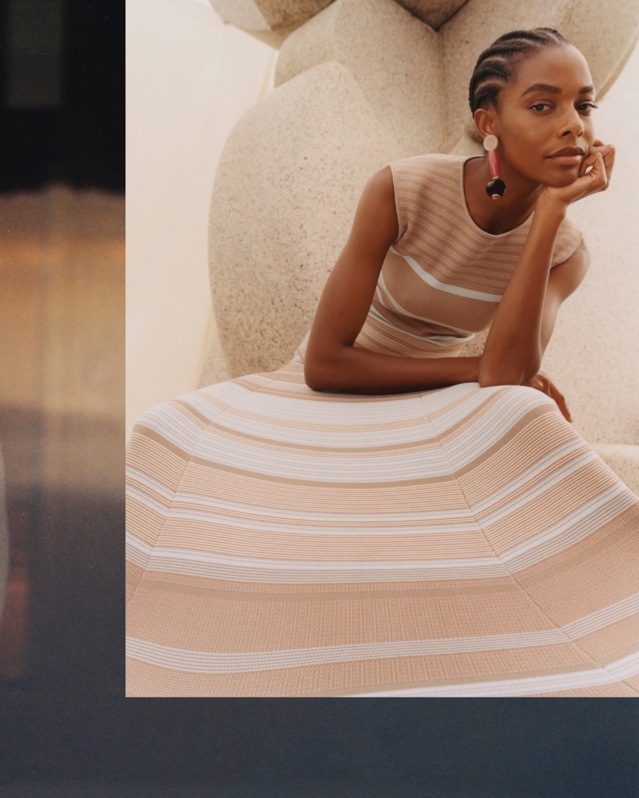 A woman sits, arms folded in thought. She wears a subtly-striped pink and sand dress from Giorgio Armani's SS22 Collection