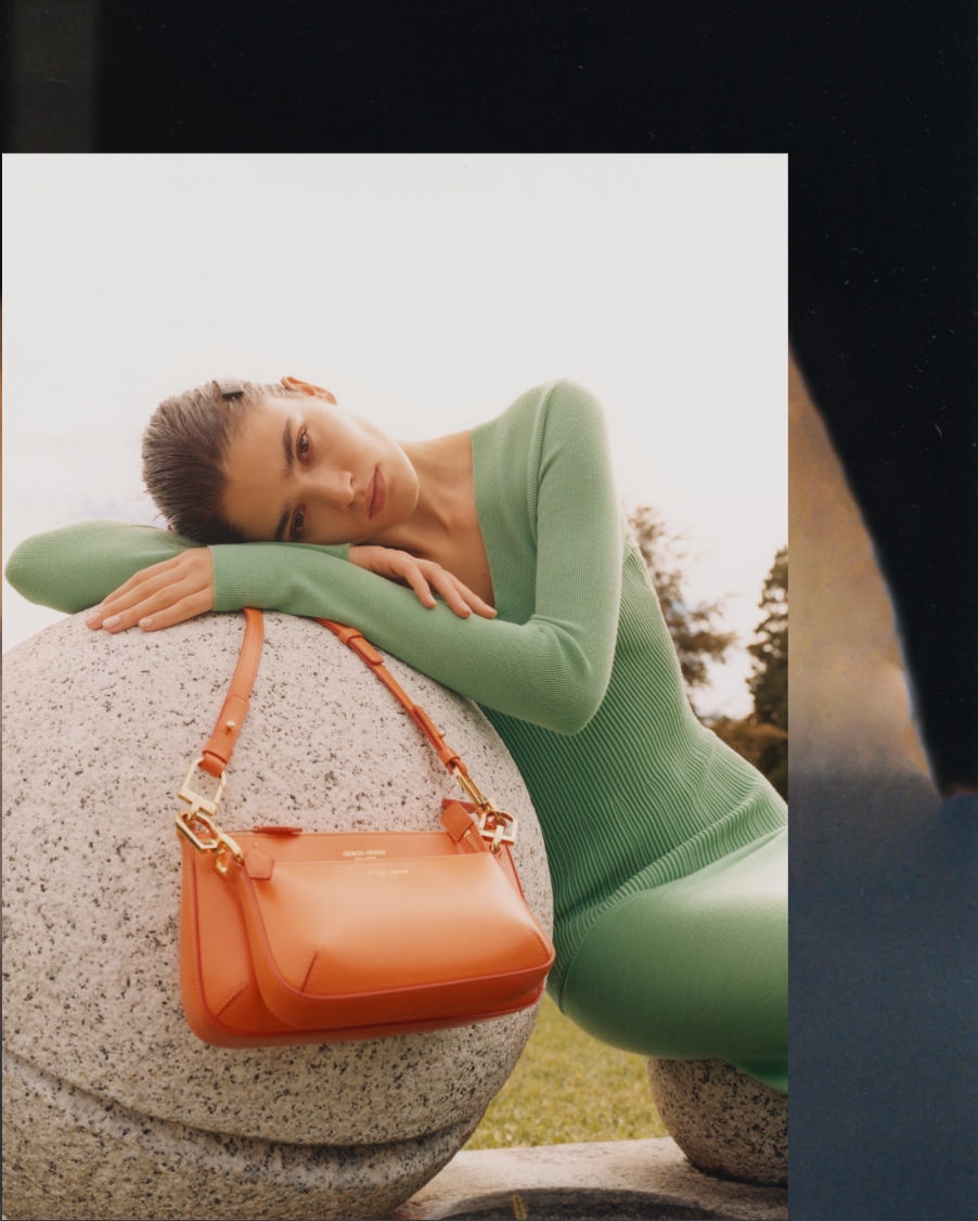 A woman in a green dress rests on a stone orb, an orange purse from Giorgio Armani's SS22 Collection draped over the side.
