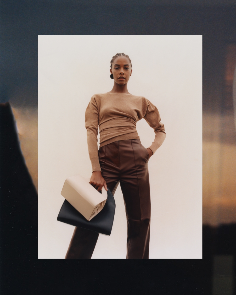 A woman wearing a brown sweater and pants from Giorgio Armani's SS22 Collection poses with two purses