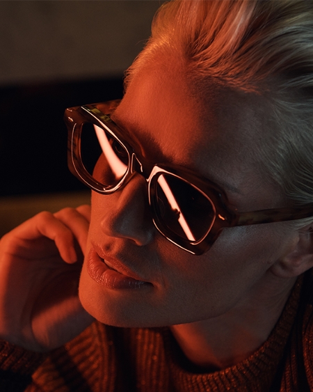 A woman with short blonde hair wears thick-framed glasses. She is lit with moody red light.