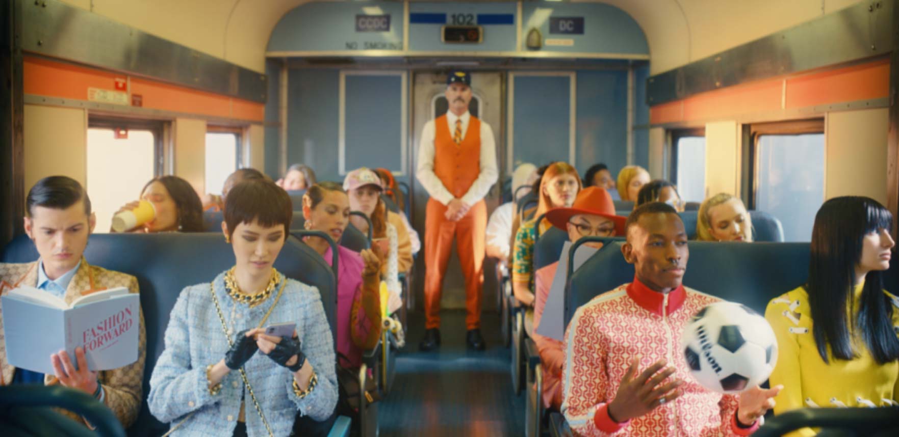 Passengers sit on a train as a conductor in an orange suit stands in the background